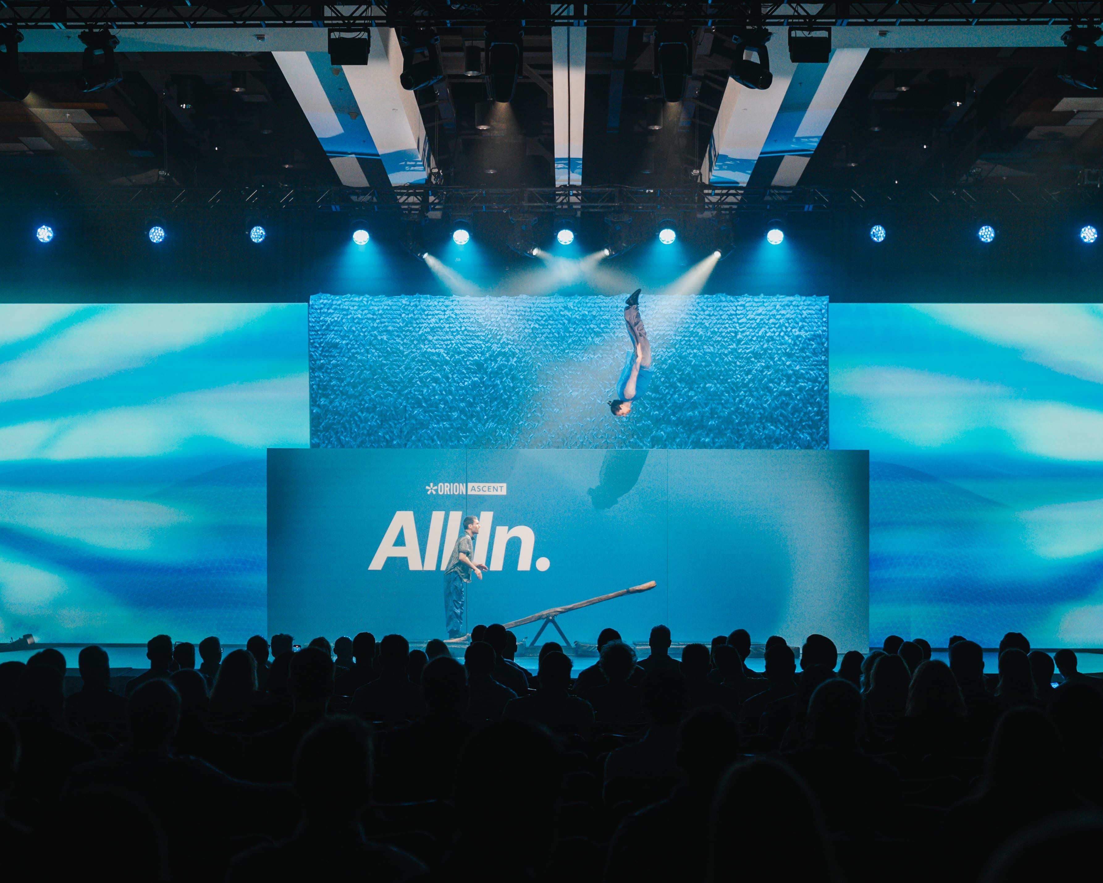 Circus acrobat flipping high above the Orion Ascent All In stage under blue lights