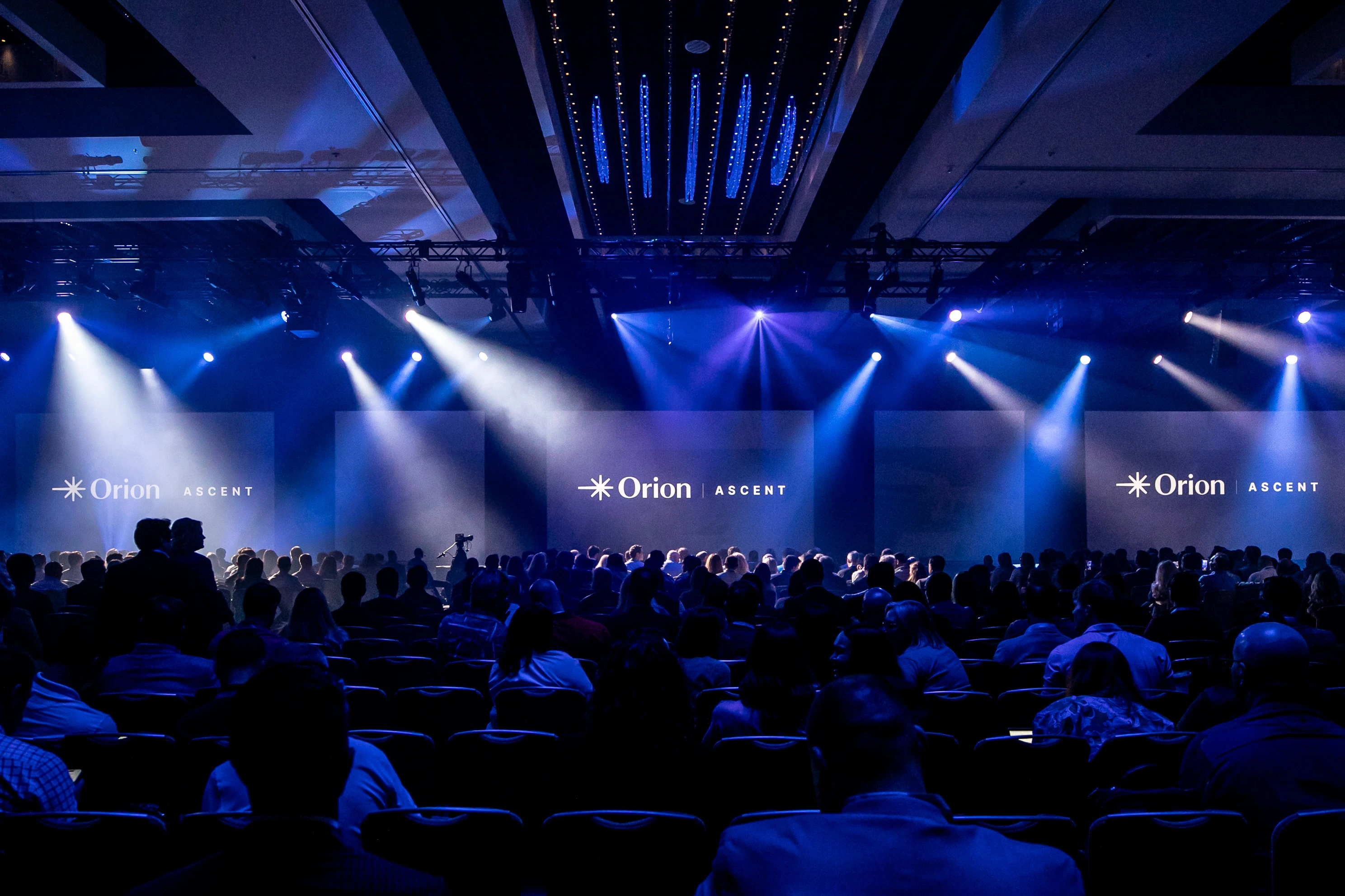 Orion Ascent logos across three screens above a blue-lit ballroom audience