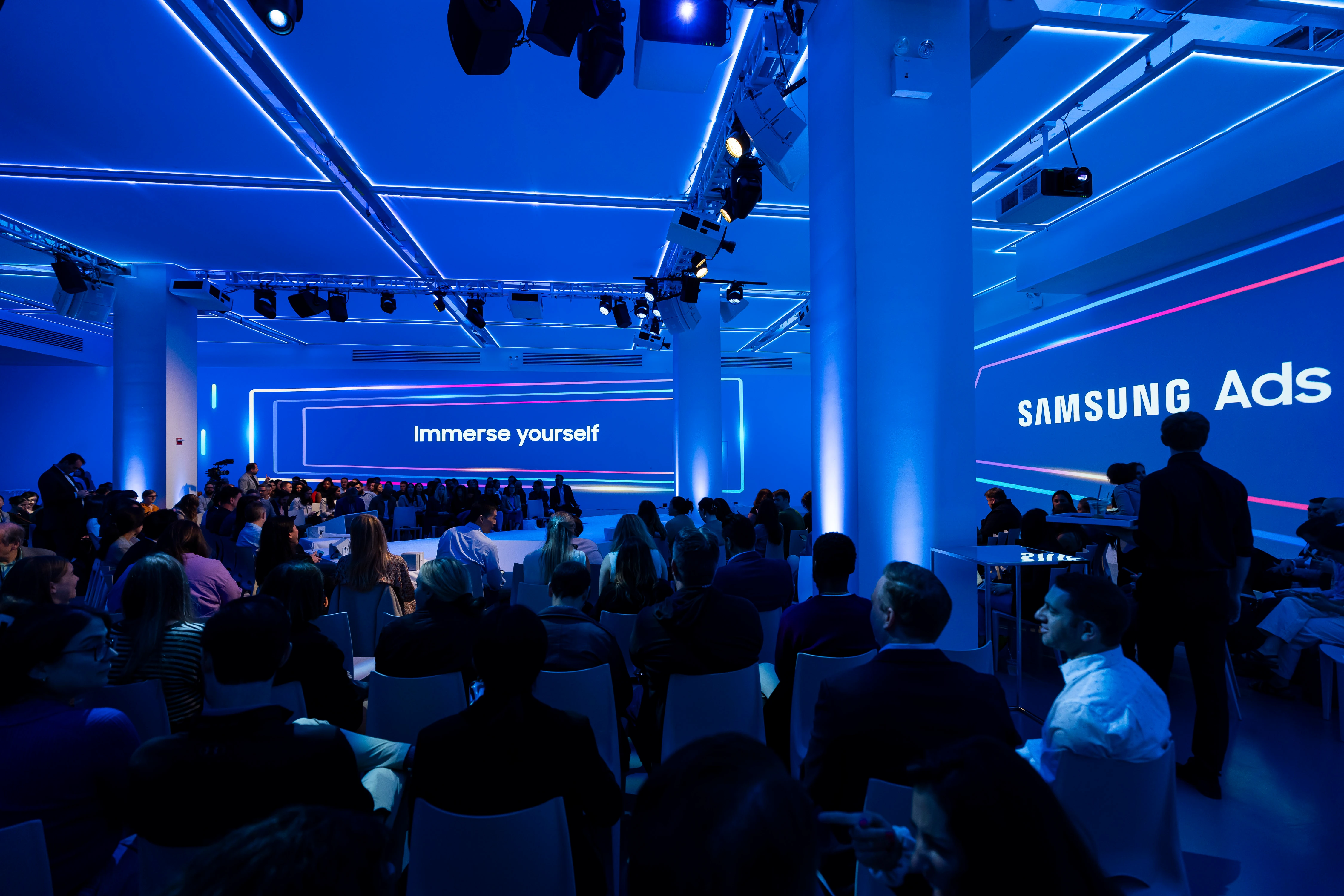 Blue-lit Samsung Newfront audience facing an Immerse Yourself screen beside a Samsung Ads wall