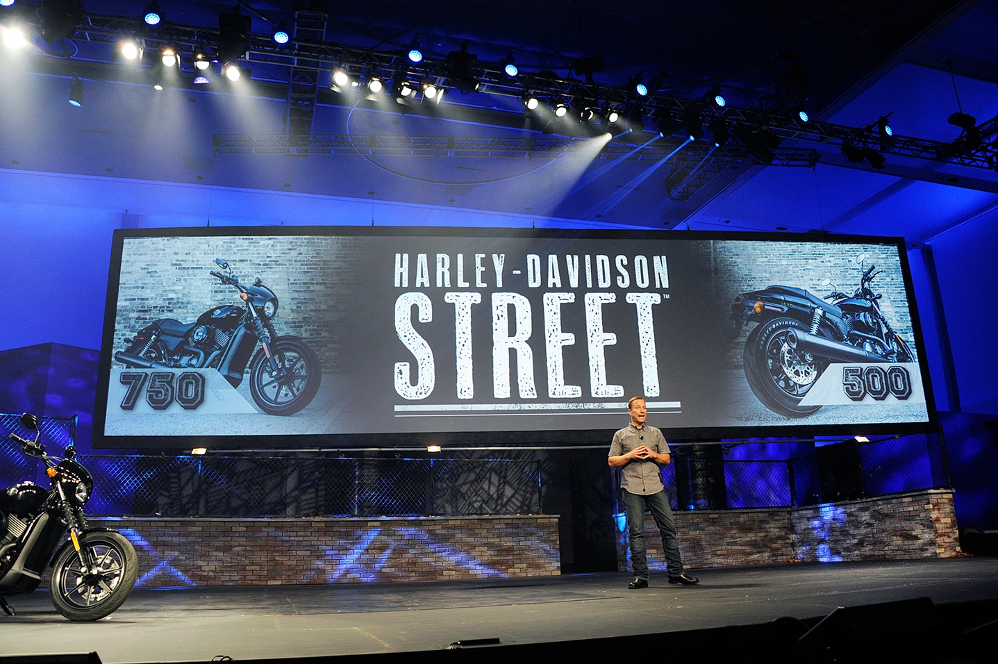 Speaker on stage beside a motorcycle under a huge Harley-Davidson Street 750 and 500 screen