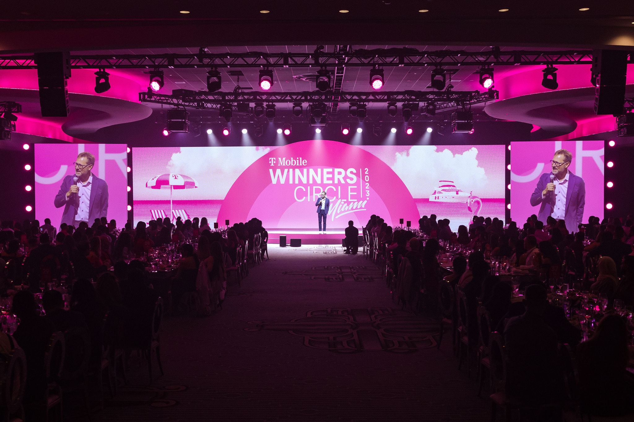 Speaker on the T-Mobile Winners Circle Miami 2023 stage between dinner tables and side screens